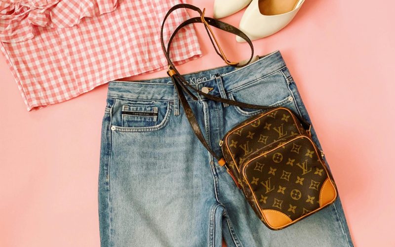 A trendy flat lay of a chic summer outfit featuring denim jeans, crop top, heels, and a stylish bag on a pink background.