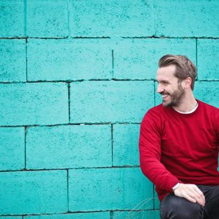 A cheerful man in a red sweater sits against a turquoise wall, enjoying a moment of happiness and laughter.
