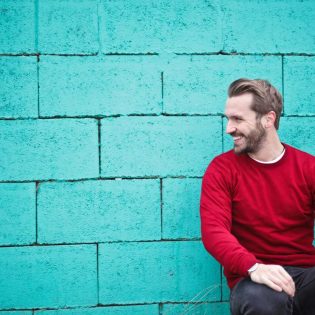 A cheerful man in a red sweater sits against a turquoise wall, enjoying a moment of happiness and laughter.